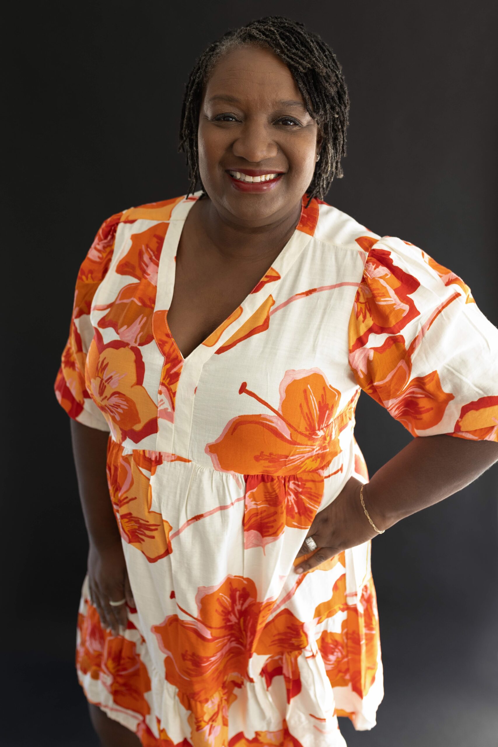 K Paige Engle smiling in a orange and white flower dress with hand on hip